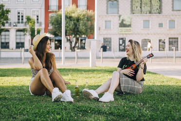 Cheerful woman playing ukulele while sitting with female friend on grassy land - DCRF00792