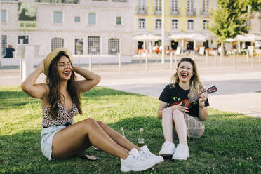 Carefree woman playing ukulele while sitting with friend on grassy land - DCRF00793