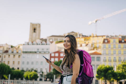 Happy woman with backpack using smart phone while standing against buildings in city stock photo