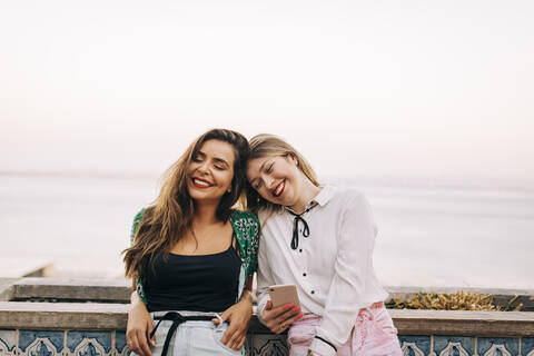 Smiling female friends with eyes closed sitting against sea stock photo