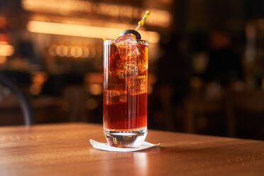 Highball-Glas mit rotem Alkohol-Cocktail mit Eiswürfeln, dekoriert mit einem Stock mit schwarzer Olive auf einem Holztisch - ADSF14806