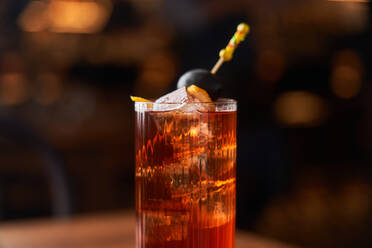 Highball-Glas mit rotem Alkohol-Cocktail mit Eiswürfeln, dekoriert mit einem Stock mit schwarzer Olive auf einem Holztisch - ADSF14807