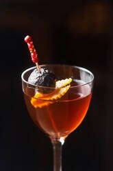 Roter Alkohol-Cocktail Manhattan garniert mit Kirsche und Orangenschale am Stiel vor dunklem Hintergrund - ADSF14810