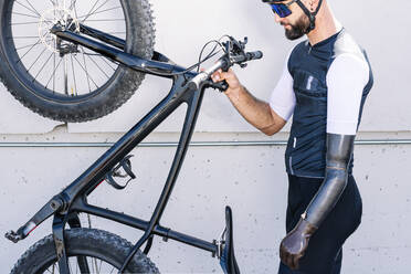 Male amputee athlete holding bicycle while standing by wall - JCMF01253