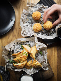 From above top view cropped unrecognizable person hand holding delicious deep fried cheese balls near plate with dumplings garnished with green herbs placed on paper served with white sauce on wooden table - ADSF14852