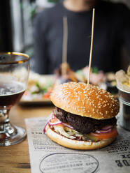 Appetitliche Rindfleisch-Burger mit Tomaten und Zwiebeln mit Weizenbrötchen mit Sesam serviert auf Papier auf Restaurant Tisch mit Ernte unerkennbar Client im Hintergrund - ADSF14854