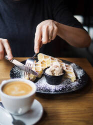 Unerkennbare Person mit Form und Messer zu schneiden süße Waffeln in der Nähe Schüssel Eis und Tasse Kaffee auf dem Tisch im Café - ADSF14860