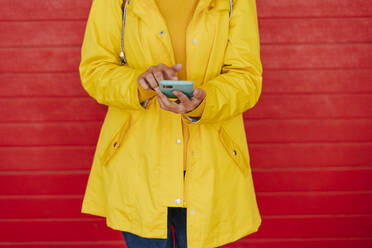 Frau im gelben Regenmantel mit Smartphone vor roter Wand - EBBF00685
