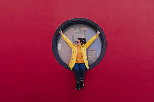 Woman in yellow rain coat sitting in porthole making victory sign - EBBF00688