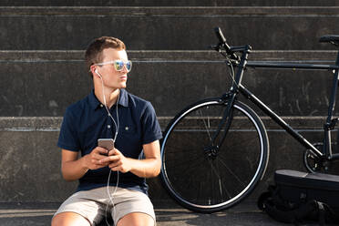 Junger Mann, der mit seinem Fahrrad auf einer Betontreppe sitzend ein Mobiltelefon benutzt - ADSF14871