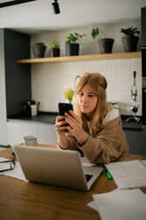 Side view of pensive female accountant sitting at table using on mobile phone while having break during remote work at home and laughing - ADSF14872