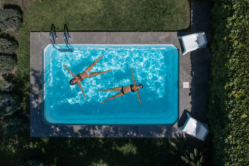 From above drone view of relaxed young female friends lying on water in pool and enjoying summer vacation while spending time together in green yard - ADSF14889