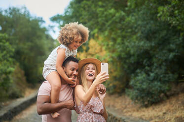 Cheerful multiethnic parents with cute curly hair daughter having fun and taking selfie on smartphone while spending summer day together in park - ADSF14896