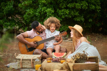 Glückliche junge gemischte Rasse Eltern mit niedlichen lockiges Haar Tochter Gitarre spielen und lachen beim Chillen zusammen während Picknick im Sommer Tag im grünen Wald - ADSF14899