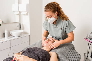 Professional beautician doctor in protective mask applying facial cream on woman while preparing for dermatology procedure in modern salon - ADSF14901