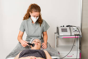 Relaxed female client getting skincare facial massage with ultrasound device from professional cosmetician in protective face mask in contemporary beauty clinic - ADSF14903