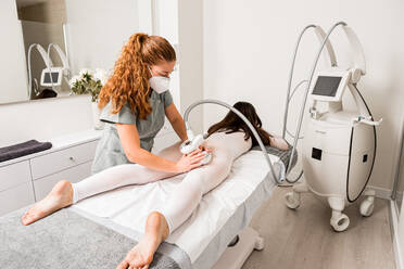 Masseuse in protective mask doing anti cellulite lipo massage to female client dressed in white suit during body care procedure in salon of apparatus cosmetology - ADSF14904