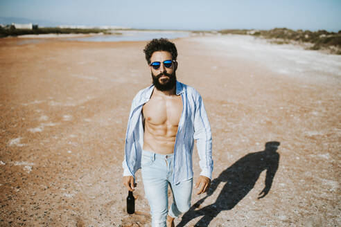 Young man with open shirt walking in desert while holding beer bottle in hand on sunny day - MIMFF00174