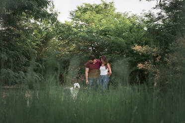 Zartes glückliches Paar, das sich umarmt, während es mit einem alten englischen Schäferhund im Garten an einem sonnigen Tag steht - ADSF14910