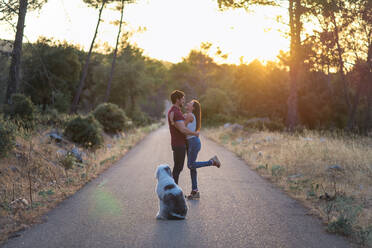 Side view of cheerful girlfriend and boyfriend standing on roadway with Old English Sheepdog and enjoying amazing sunset in summer - ADSF14913