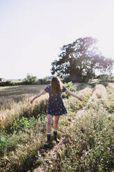 Carefree female in dress and rubber boots walking along field while enjoying weekend in countryside on sunny day - ADSF14941