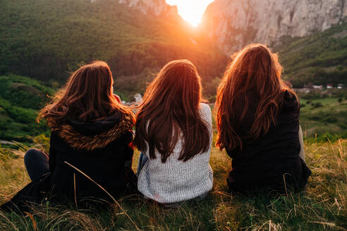 Rückenansicht von anonymen Freundinnen, die während eines malerischen Sonnenuntergangs in Transsilvanien auf einer Wiese in den Bergen chillen und über die Schulter schauen - ADSF14948