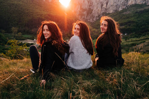 Rückenansicht von charmanten Freundinnen, die während eines malerischen Sonnenuntergangs in Siebenbürgen auf einer Wiese in den Bergen chillen und über die Schulter schauen - ADSF14949