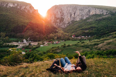 Group of friendly people relaxing on lawn in highlands at sunset and taking selfie on smartphone during vacation in Transylvania - ADSF14950