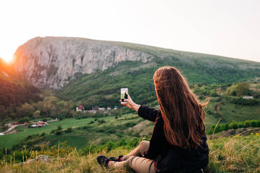 Back view of unrecognizable female tourist sitting on grass and taking selfie on mobile phone camera while resting in highlands during summer holiday in Transylvania - ADSF14951