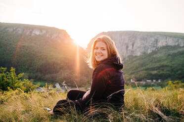 Back view of cheerful female tourist sitting on grass resting in highlands during summer holiday in Transylvania - ADSF14953