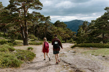 Rückenansicht eines nicht erkennbaren jungen Paares von Reisenden mit Rucksäcken, die auf einem Pfad zwischen grünen Wäldern und Bergen an einem Sommertag in Navacerrada in Spanien wandern - ADSF14957