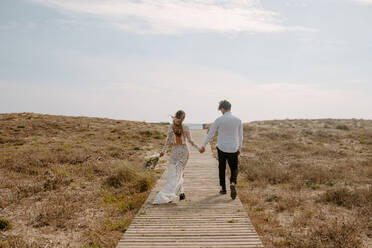 Full body back view of unrecognizable young newlywed couple in stylish clothes with bouquet holding hands and walking on wooden pathway towards sea - ADSF14961