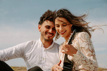 Glückliches junges frisch verheiratetes Paar in böhmischer eleganter Kleidung spielt Ukulele-Gitarre, während sie zusammen am Sandstrand sitzen - ADSF14963
