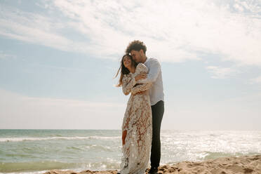 Ganzkörper-Seitenansicht von fröhlichen jungen Braut und Bräutigam in stilvollen Hochzeitskostümen stehen umarmt einander mit geschlossenen Augen auf Sandstrand in der Nähe winken Meer und genießen einander - ADSF14964