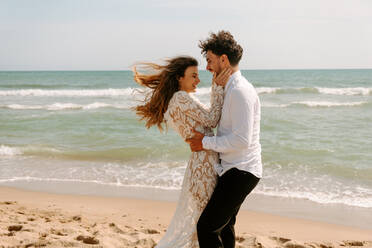Seitenansicht von fröhlichen jungen Braut und Bräutigam in stilvollen Hochzeitskostümen stehen umarmt einander mit geschlossenen Augen auf Sandstrand in der Nähe winken Meer und genießen einander - ADSF14965