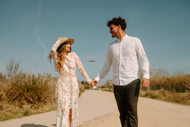 Glücklicher junger Mann in stilvollem Outfit, der die Hand einer Frau im weißen Hochzeitskleid hält, während sie gemeinsam auf einer Landstraße gegen den blauen Himmel mit einem fliegenden Flugzeug spazieren gehen - ADSF14967
