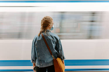 Rückenansicht einer weiblichen Passagierin in Schutzmaske und lässiger Jeansjacke mit Handtasche, die auf einem U-Bahnsteig vor einem fahrenden Zug steht - ADSF14970