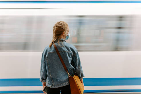 Back view of millennial female passenger in protective mask and casual denim jacket with handbag standing on underground platform in front of moving train - ADSF14970