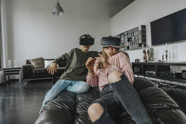 Excited friends looking through virtual reality simulators while relaxing on couch at home - MFF06096