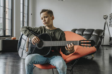 Junge spielt Gitarre, während er auf einem Stuhl zu Hause sitzt - MFF06113