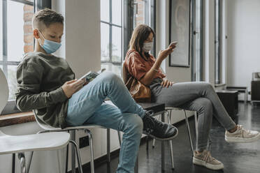 Boy and teenage girl wearing masks sitting on chairs in waiting room - MFF06118