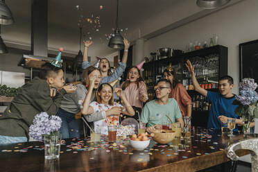 Cheerful friends throwing confetti on teenage girl in birthday party at home - MFF06122