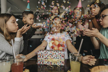 Happy friends throwing confetti on teenage girl sitting with birthday gift at dining table - MFF06125