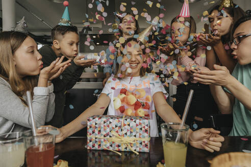 Happy friends throwing confetti on teenage girl sitting with birthday gift at dining table - MFF06125