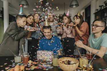 Friends throwing confetti on birthday boy sitting with gift at dining table - MFF06126