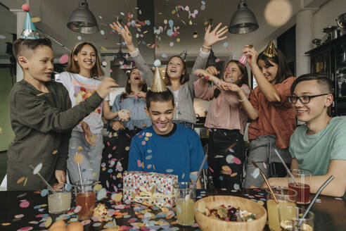 Cheerful friends throwing confetti on birthday boy sitting with gift at dining table - MFF06127