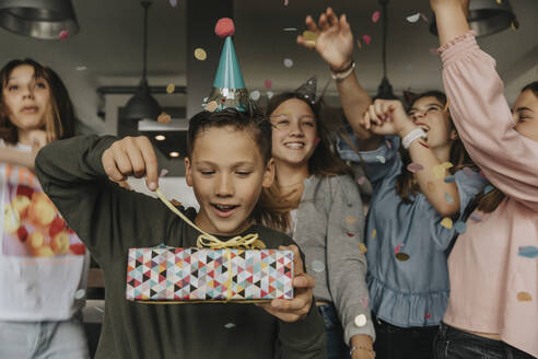 Curious birthday boy opening gift while friends dancing in background - MFF06131