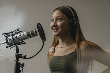 Close-up of smiling teenage girl singing over microphone against wall in recording studio - MFF06175