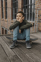 Lächelnder Junge zeigt Friedenszeichen, während er auf einem Skateboard vor einem Gebäude sitzt - MFF06187