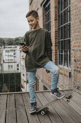 Junge benutzt sein Smartphone, während er mit seinem Skateboard auf der Promenade gegen das Geländer fährt - MFF06190